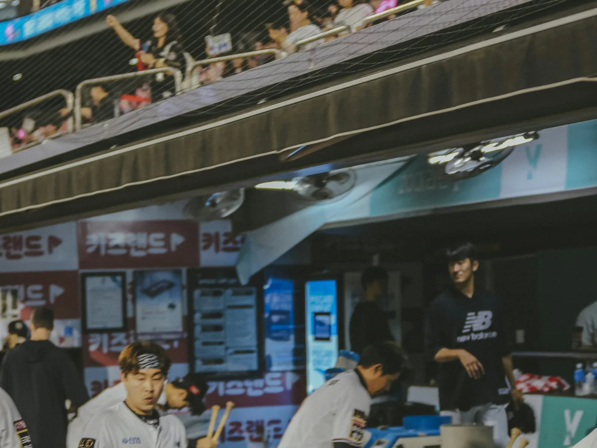 KBO-spillere nær dugout med fans presset mod nettet ovenover, repræsentativt for publikumsenergien på et koreansk baseballstadion